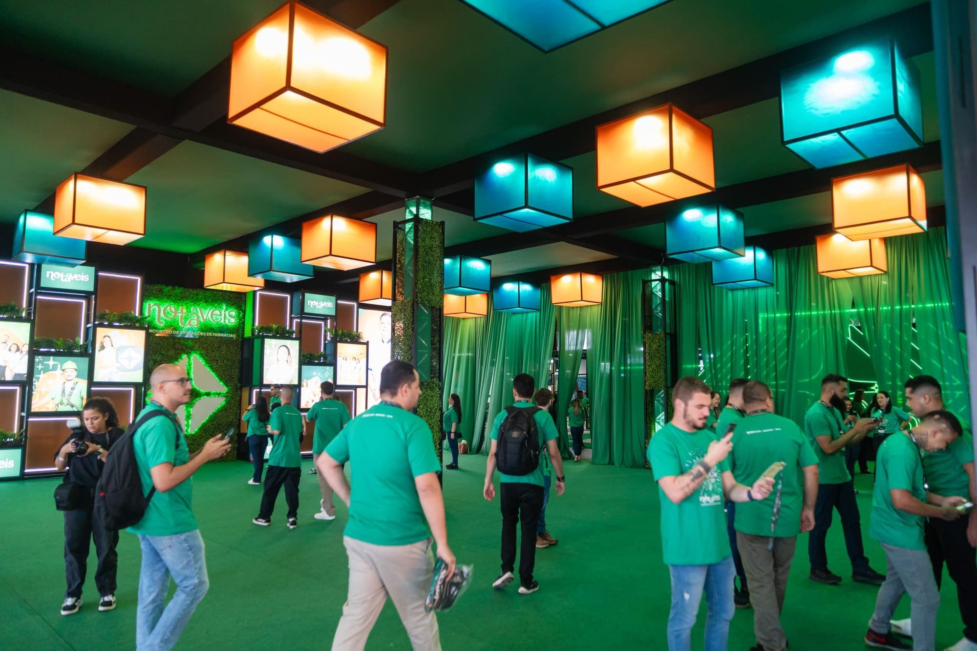Área de convivência com luminárias cúbicas coloridas suspensas, cortinas verdes e participantes circulando