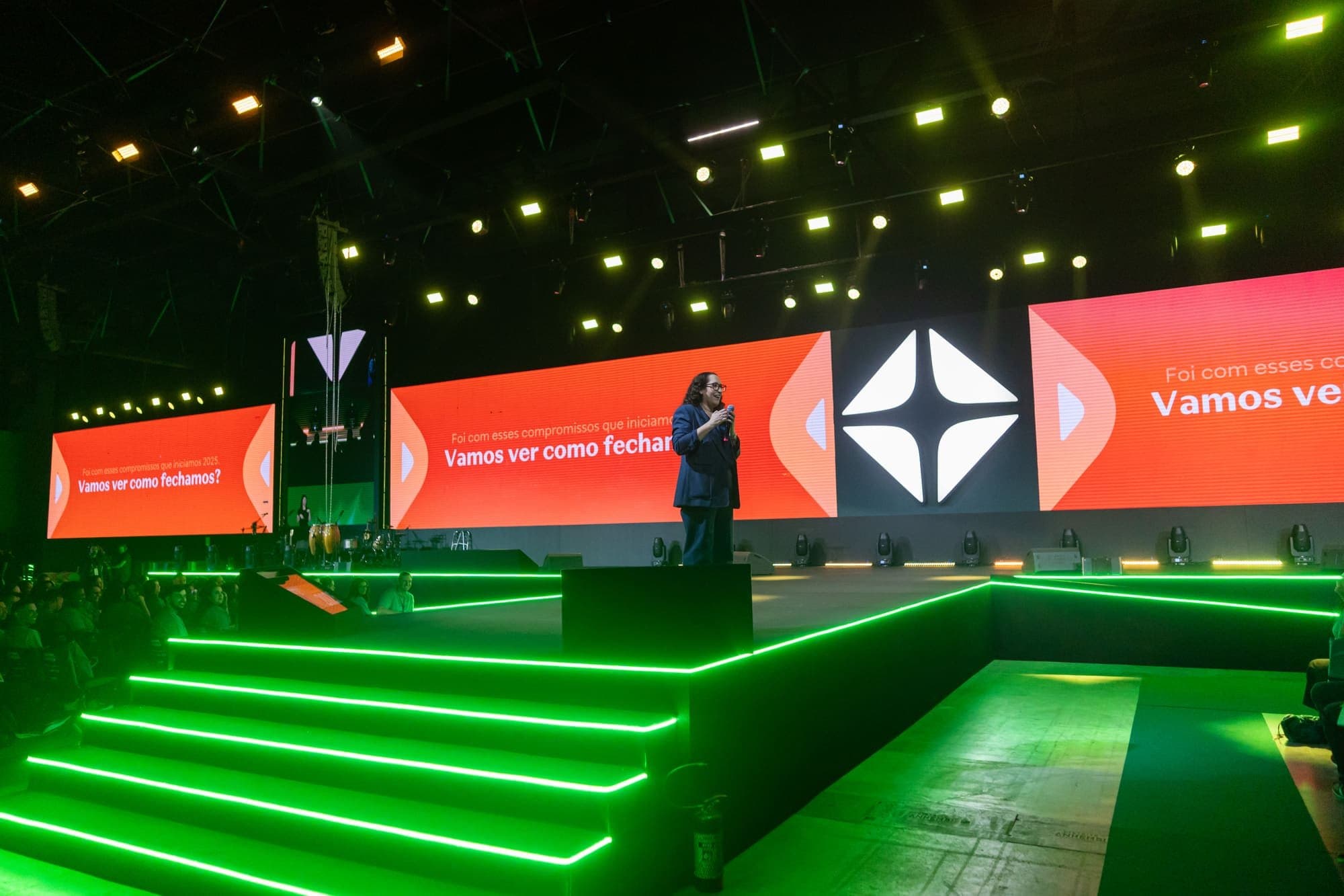Palco com degraus contornados por fitas de LED verde e três painéis de LED ao fundo, com uma apresentadora falando ao microfone