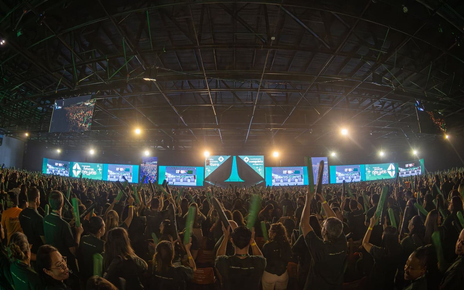 Plenária lotada diante de um telão de LED que percorre toda a largura do palco, sob a estrutura de truss do pavilhão