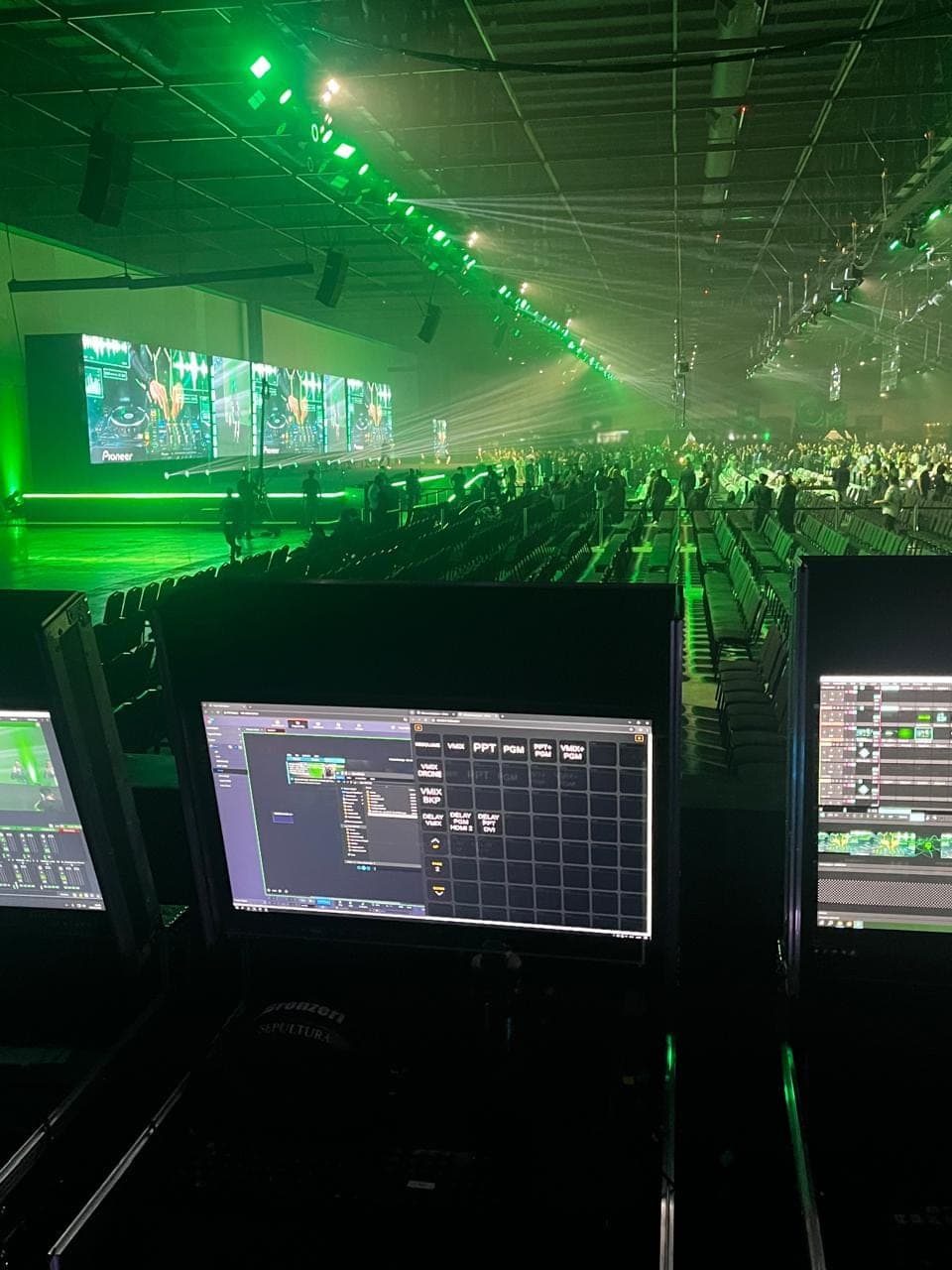 Posição de operação com multiview e monitores em primeiro plano, diante de uma arena com telões de LED, feixes de luz e plateia cheia