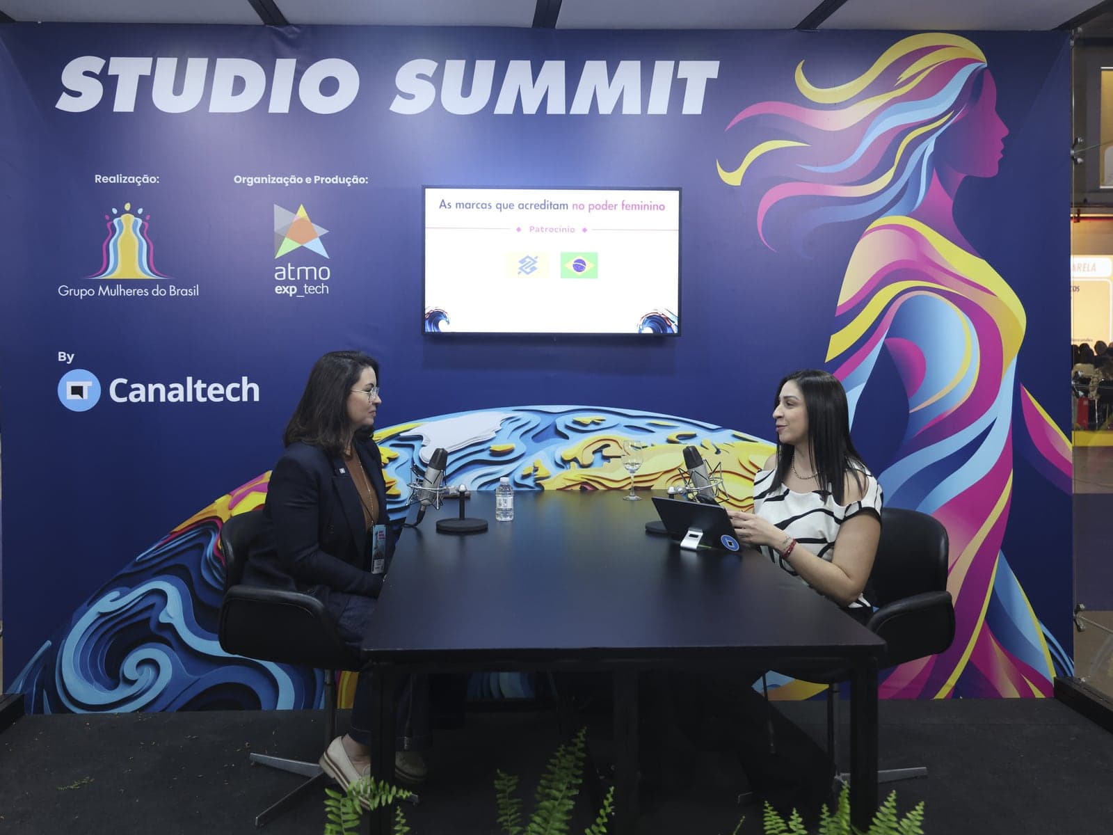 Estúdio de podcast montado pela Atmo dentro do Summit Mulheres nas Profissões, visto de ponta a ponta, com cenografia, tela e duas participantes à mesa